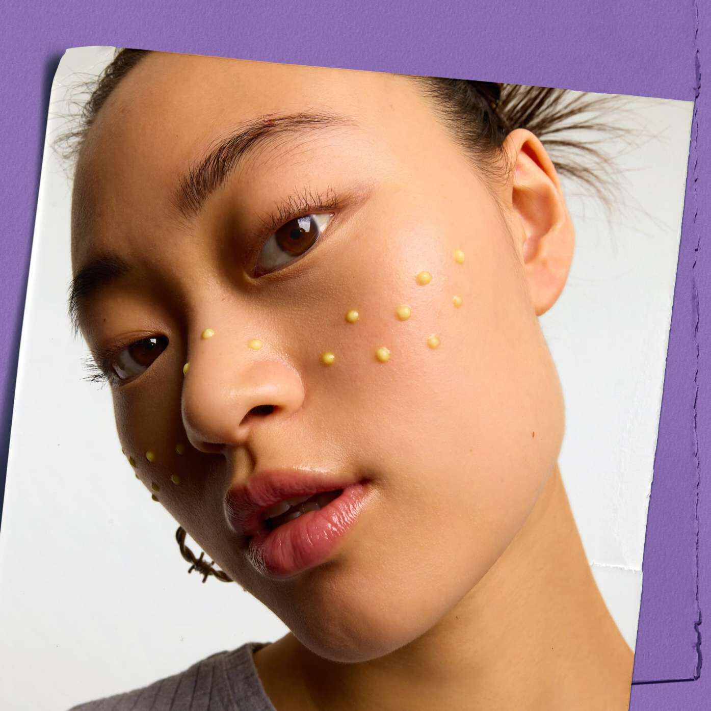 Close-up of a person with a serene expression, facing the camera. Small, yellow dots are applied in a symmetrical pattern across the cheeks. Purple background.