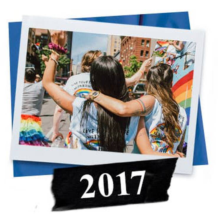 Two women embrace and celebrate at a colorful Pride event in 2017, wearing matching shirts and surrounded by vibrant rainbow decorations.