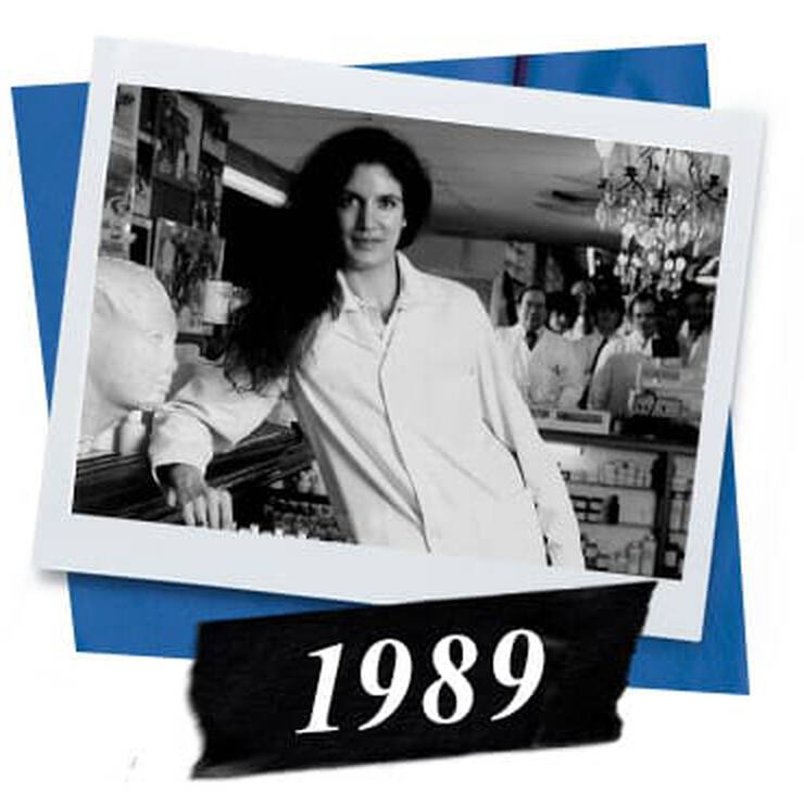 A vintage black and white photo from 1989 featuring a person in a white shirt leaning against a counter with shelves full of items behind.