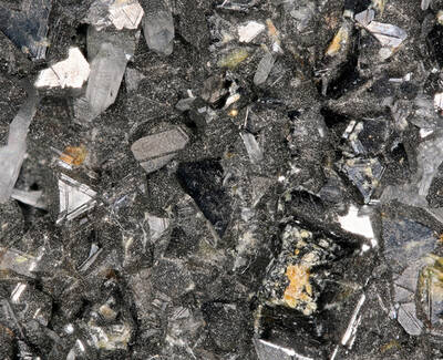 A close-up image of a cluster of dark, shiny mineral crystals with various textures and shapes, set against a backdrop of smaller sparkling particles.
