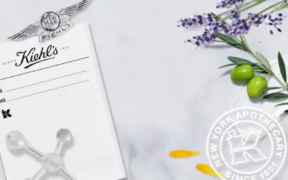 Alt text: A collage featuring a historic black-and-white photo of Kiehl's pharmacy staff, surrounded by a marble background with lavender, olive leaves, and decorative elements, showcasing the brand's heritage since 1851.