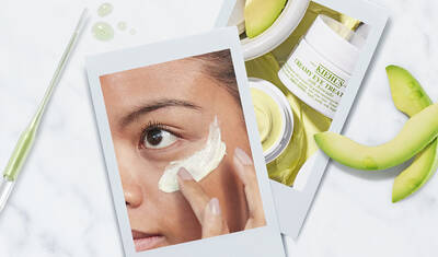 A close-up of a person applying cream under their eye, with beauty products displayed nearby, including a creamy eye treatment and slices of green fruit.