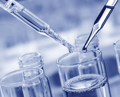 Two pipettes are poised above glass test tubes containing clear liquid, with water droplets ready to be released. The image has a cool blue tint.
