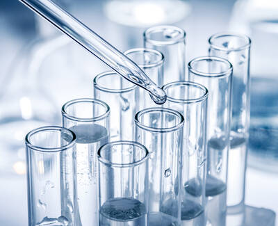 A close-up of several glass test tubes filled with clear liquids, with a dropper releasing a drop of liquid into one of the tubes.