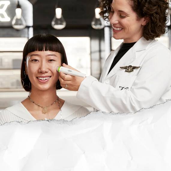 A smiling woman with a bob haircut is receiving a facial treatment from a technician in a lab coat, who is using a device on her cheek. The setting has a modern, bright aesthetic with pendant lights in the background.