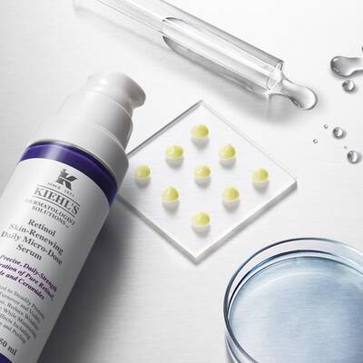 A flat lay image featuring a bottle of skin-renewing retinol serum with a purple label, accompanied by a glass dropper, small droplets of liquid, and several yellow drops on a clear petri dish. The background is light and reflective.