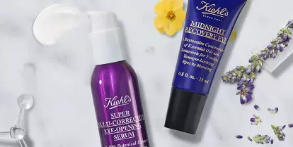 A flat lay of Kiehl's skincare products, including a green avocado eye cream, a lavender serum, and a purple moisturizer, arranged on a marble background alongside a dropper and an avocado slice.