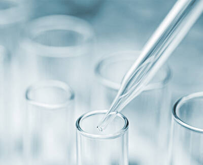 A clear pipette is poised above a glass test tube, with a blurred background of several other test tubes.