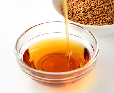 A glass bowl containing golden-brown oil, with sesame seeds in a white bowl in the background.