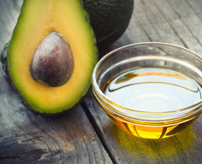 A sliced avocado revealing its pit alongside a small bowl of golden avocado oil, placed on a wooden surface.