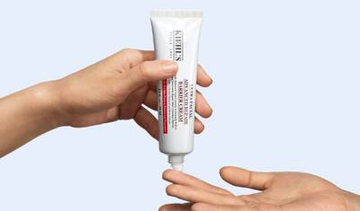 A hand is holding a tube of Kiehl's skincare cream, while another hand is reaching out to receive the product. The background is light blue.