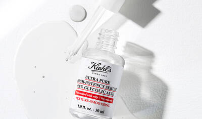 A bottle of Kiehl's Ultra Pure High-Potency Serum with 39% Glycolic Acid is lying on a white surface, with a small amount of serum spilled nearby. The dropper cap is slightly raised, showcasing the product's clean, minimalist design.
