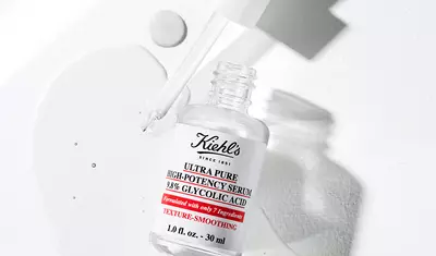 A bottle of Kiehl's Ultra Pure High-Potency Serum with 39% Glycolic Acid is lying on a white surface, with a small amount of serum spilled nearby. The dropper cap is slightly raised, showcasing the product's clean, minimalist design.