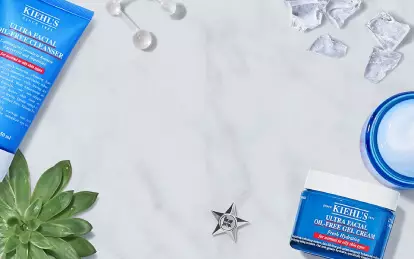 A flat lay of Kiehl's skincare products, including an Ultra Facial Oil-Free Cleanser, an Ultra Facial Oil-Free Toner, and a moisturizer, surrounded by ice cubes, a lemon slice, and a succulent plant on a marble surface.