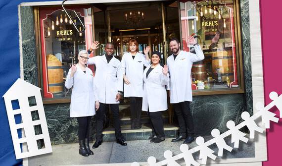 A group of six people in white lab coats stand outside a Kiehl’s store, smiling and waving, with a vintage storefront in the background.