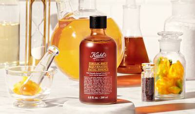 A bottle of Kiehl's Ferulic Brew Rejuvenating Facial Essence surrounded by various glass containers filled with liquids and colorful ingredients, set on a light background.