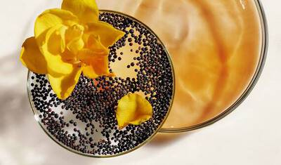 A close-up of two glass dishes: one filled with a light brown liquid and the other with small black seeds, topped with yellow flower petals.