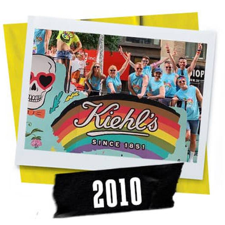 A vibrant group of people in matching blue shirts celebrates on a float decorated with Kiehl's logo and a colorful rainbow at a parade in 2010.
