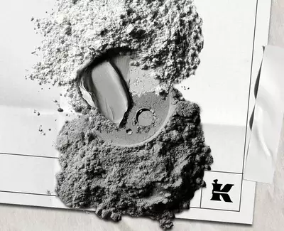 A close-up of various shades of gray powders and a cream-like texture on a crumpled white surface, featuring a stylized 'K' logo.