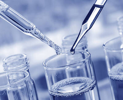 Close-up of two pipettes dispensing liquid into clear glass test tubes with bubbles in the solution. The image has a blue tint.