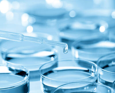 A pipette is poised above multiple petri dishes filled with a blue liquid, with a drop about to fall from the pipette.
