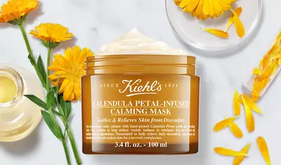 A jar of Kiehl's Calendula Petal-Infused Calming Mask sits on a marble surface, surrounded by orange calendula petals and a small bottle of oil. The jar is labeled and highlights the product's soothing and skin-relieving properties.