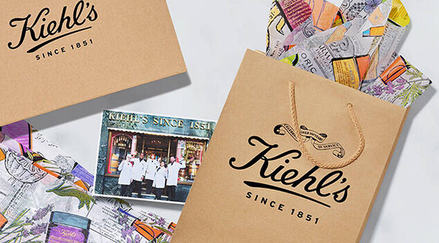 Three Kiehl's gift boxes with a beige design and white ribbons are displayed against a marbled background, alongside colorful artistic wrapping paper.