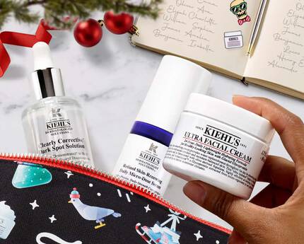 A festive flat lay featuring a zipped cosmetic pouch with winter-themed designs, containing three skincare products: a serum, a moisturizer, and a facial cream, alongside a lit candle and holiday decorations.