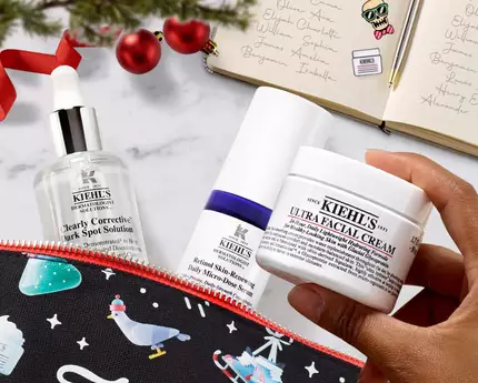 A festive flat lay featuring a zipped cosmetic pouch with winter-themed designs, containing three skincare products: a serum, a moisturizer, and a facial cream, alongside a lit candle and holiday decorations.