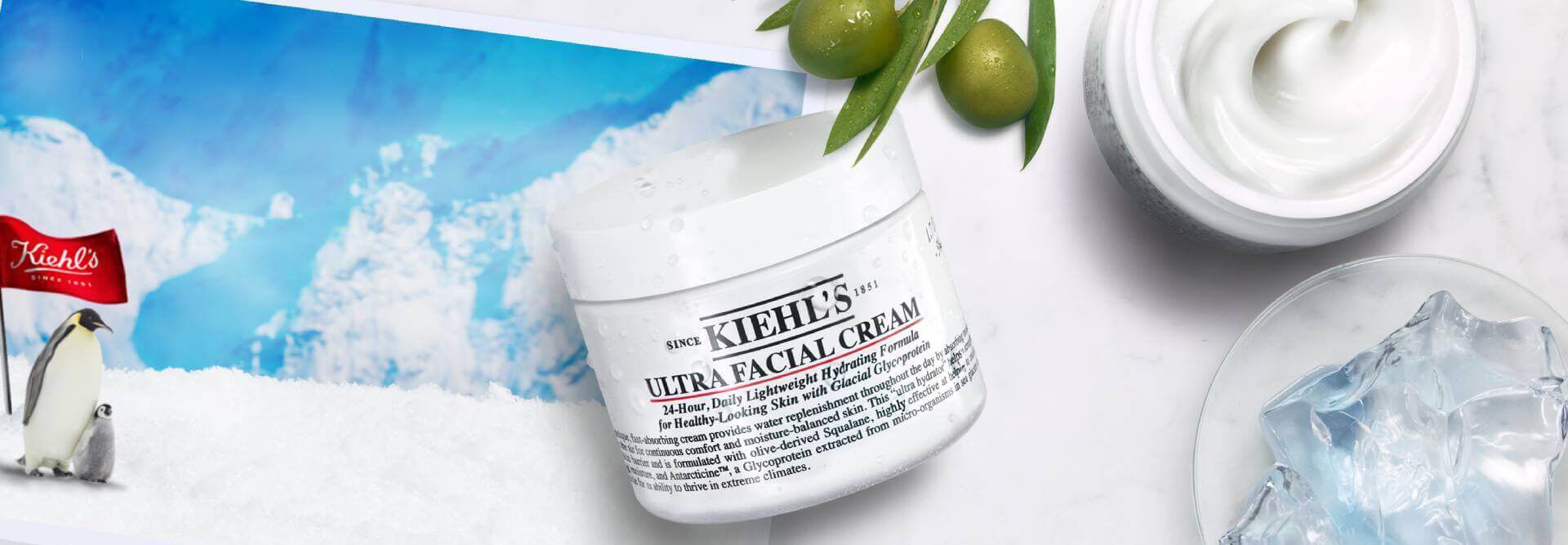 A jar of Kiehl's Ultra Facial Cream is displayed against a backdrop of icy mountains and snow, accompanied by olive branches and a small bowl of cream, emphasizing hydration and freshness.