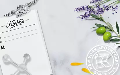 A vintage black-and-white photo of the Kiehl's pharmacy storefront, surrounded by lavender, olive branches, and yellow petals, with a white logo and text indicating the brand's history and establishment date.