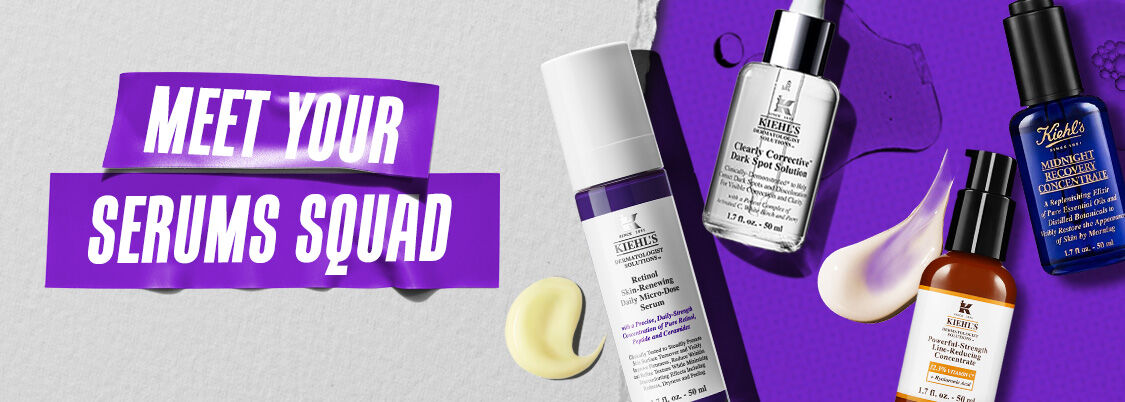 Four skincare bottles are lined up on a vibrant purple background with cream smudges, next to a textured gray area. The scene conveys a sense of luxury and care.