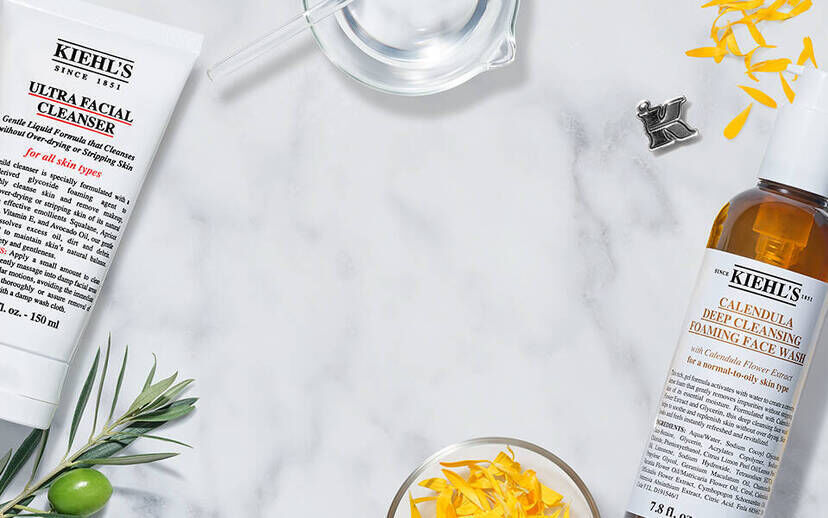 Flat lay arrangement of Kiehl's skincare products, including a facial cleanser, exfoliating scrub, and facial fuel, surrounded by natural ingredients like flowers, coffee beans, and fruits on a marble background.