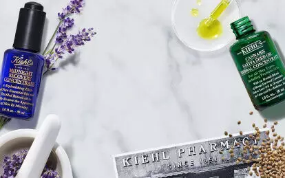 A flat lay of Kiehl's skincare products on a marble surface, featuring a blue bottle labeled "Midnight Recovery Concentrate," a green bottle labeled "Cannabis Sativa Seed Oil Herbal Concentrate," and a yellow bottle labeled "Daily Reviving Concentrate," surrounded by lavender, sunflower, and scattered seeds.