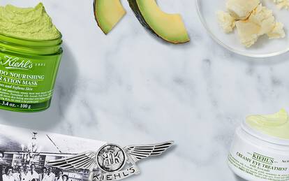 A flat lay featuring Kiehl's skincare products, including an Avocado Nourishing Hydrating Mask and a creamy treatment, surrounded by avocado slices and shea butter on a marble surface.