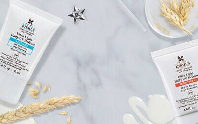 A flat lay of skincare products, including Kiehl's Ultra Light Daily UV Defense and Ultra Light Daily UV Defense for sensitive skin, along with natural ingredients like oats and transparent gel drops, all set on a marble surface.