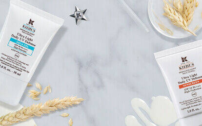 Alt text: A flat lay of Kiehl's skincare products, including Ultra Light Daily UV Defense and Ultra Light Daily UV Defense Aqua Gel, surrounded by natural elements like oats and decorative items on a marble surface.