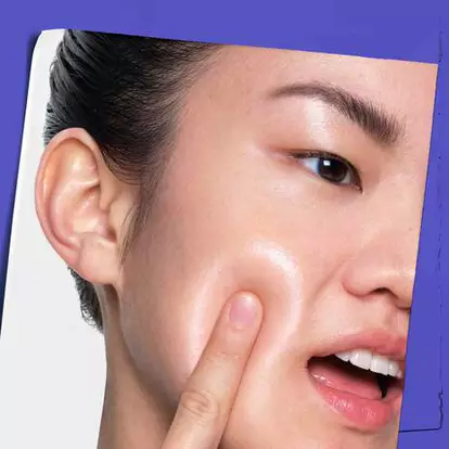 Close-up of a person's face with a focus on smooth, glowing skin. A finger touches the cheek, demonstrating a dewy, hydrated texture. The background is purple.
