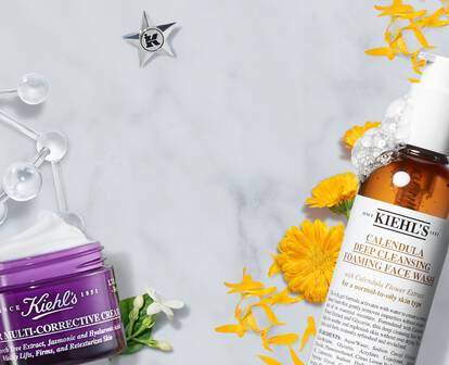 A flat lay of Kiehl's skincare products, including a brown bottle of calming serum and a purple jar of ultra-corrective cream, set against a white marble background with yellow flower accents.
