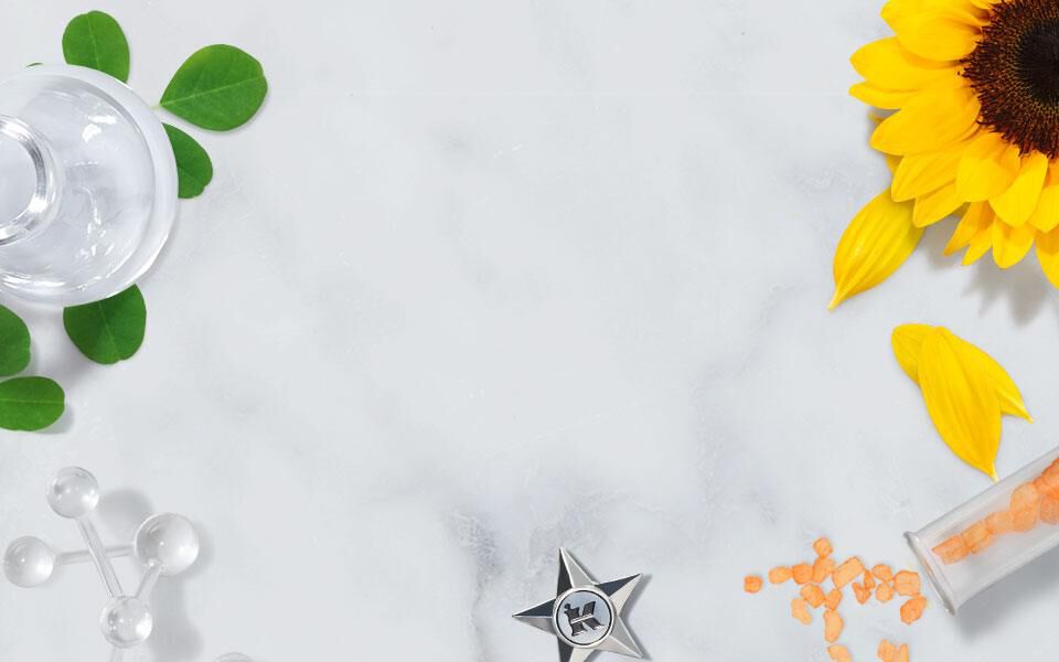 A flat lay of various natural ingredients and botanical elements, including sunflower petals, green olives, and a yellow flower, arranged on a marble surface alongside glass containers and a star-shaped ornament.